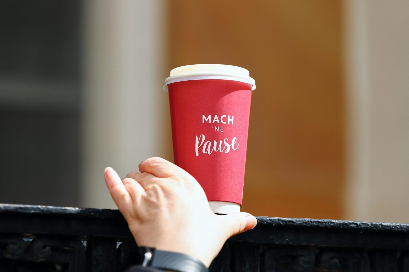 Close-up of a hand reaching for a red paper coffee cup with 'Mach ne Pause' text.